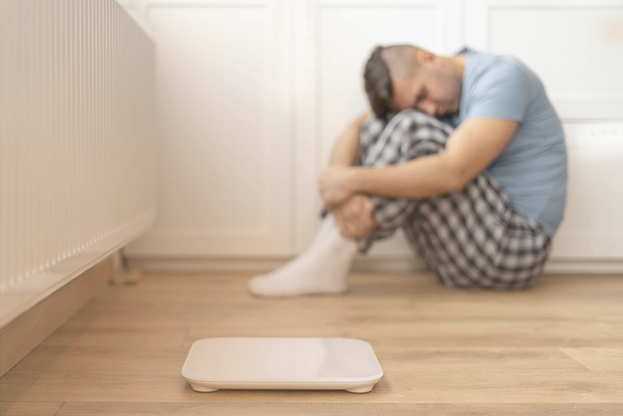 anorexia-trastorno-alimentario Un hombre en pijama sentado en el suelo abrazándose las rodillas, luciendo angustiado, con una escama blanca en primer plano, una imagen evocadora que refleja las luchas de la anorexia y otros trastornos alimentarios.