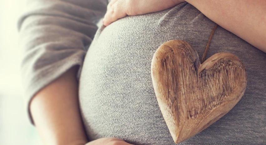 Primer plano de una persona embarazada sosteniendo suavemente su vientre, mientras un adorno de corazón de madera reposa sobre él, simbolizando el amor en medio de la ansiedad natural del embarazo.