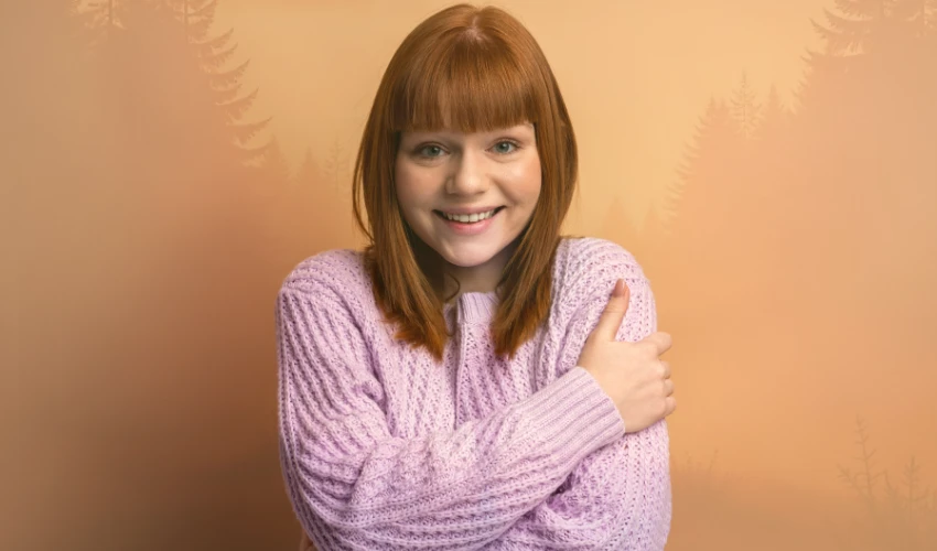 Una persona con cabello rojo y un suéter lavanda irradia confianza, sonriendo a la cámara contra un fondo beige con tenues siluetas de árboles, irradiando autoestima y valor.