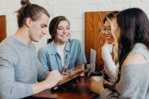 Cuatro jóvenes adultos se sientan en una mesa de café, conversando y sonriendo sobre el estrés en los estudiantes universitarios. Uno sostiene una tableta, otro una laptop y dos sostienen tazas de café mientras comparten sus experiencias y se apoyan mutuamente.