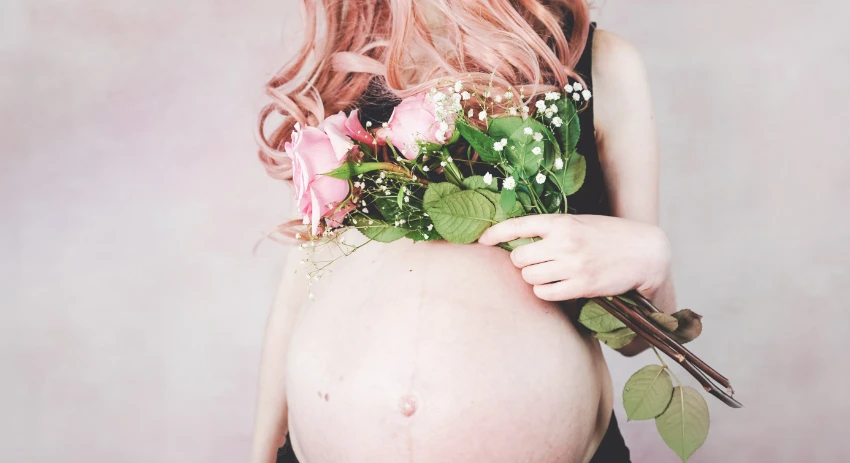 Una persona con cabello rosado sostiene rosas rosadas sobre su vientre embarazado, vistiendo de negro sobre un fondo neutro suave, capturando un momento sereno en medio de la ansiedad del embarazo.
