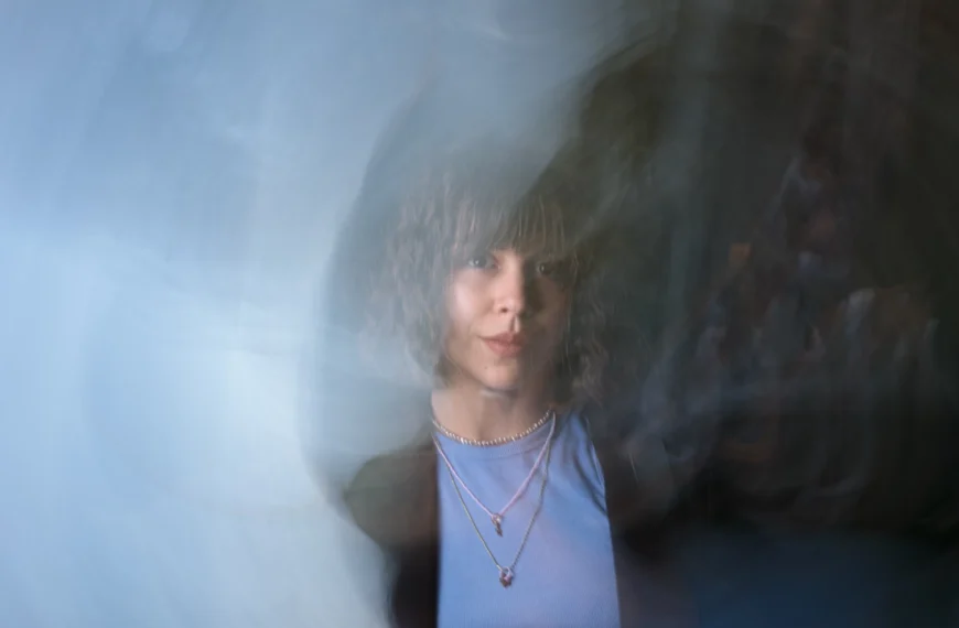 Una persona con cabello rizado y collares en capas, que encarna la autoestima como clave del éxito personal, se ve a través de un efecto de desenfoque semitransparente y giratorio mientras viste una camiseta azul.