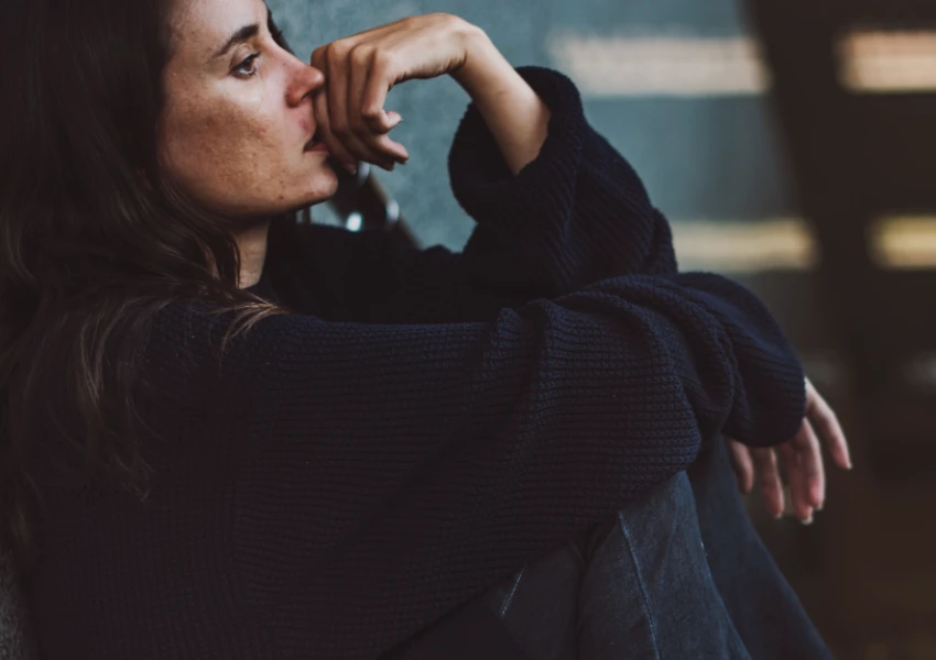 Una persona con un suéter oscuro está sentada contra una pared, pensativa, con la mano apoyada en la barbilla, como si estuviera contemplando los matices de la autoestima alta y baja.