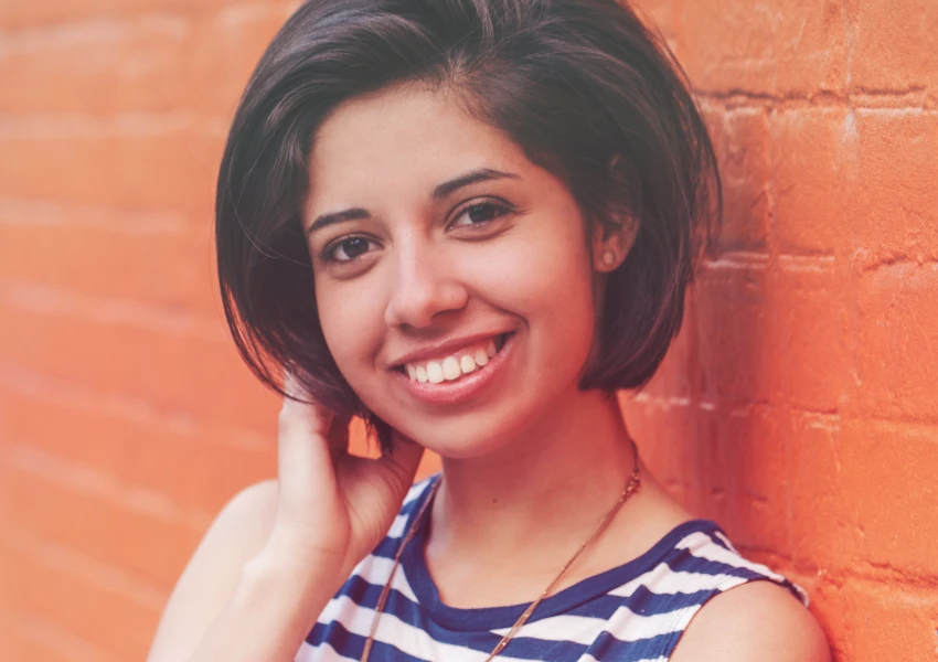 Una persona con cabello corto y oscuro sonríe con confianza, vistiendo una camiseta de rayas, mientras está parada frente a una pared de ladrillos de color naranja, exudando una sensación de alta autoestima.