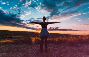 Una persona con los brazos extendidos se encuentra frente a una colorida puesta de sol, abrazada por el cielo dramático y el paisaje abierto, encarnando la relajación consciente.