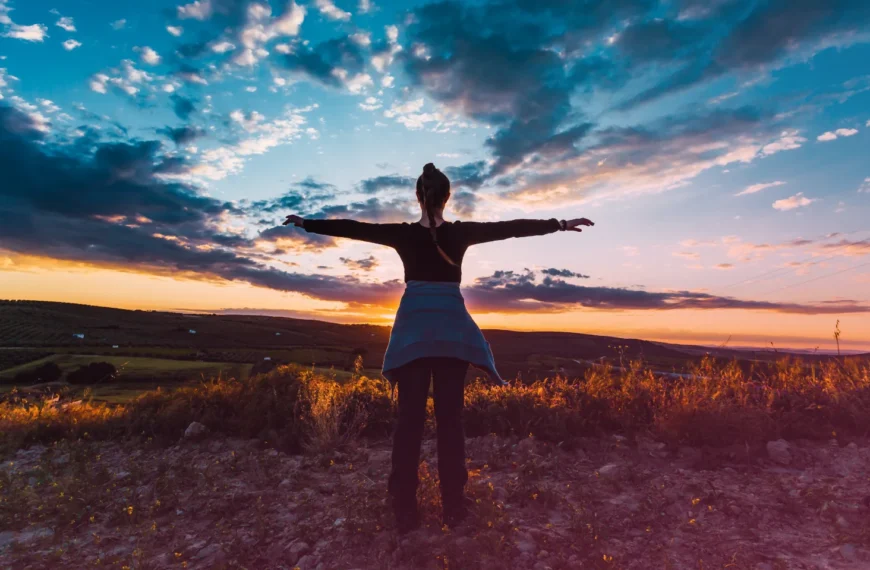 Una persona con los brazos extendidos se encuentra frente a una colorida puesta de sol, abrazada por el cielo dramático y el paisaje abierto, encarnando la relajación consciente.