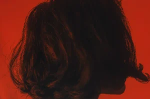 Se muestra a una persona con cabello corto y oscuro de espaldas y de lado sobre un fondo rojo, capturando la ansiedad que se siente en un momento de tensión tranquila.