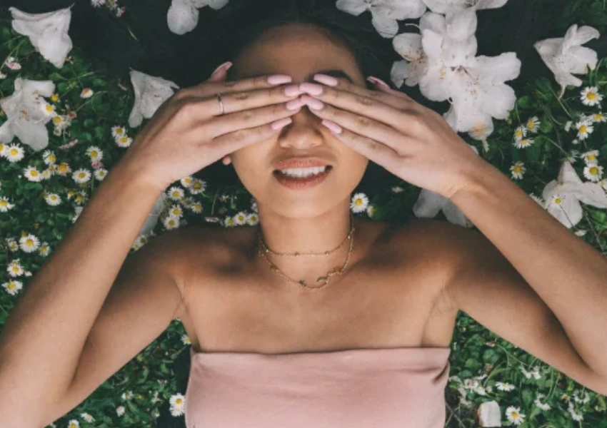 Mujer acostada sobre césped y flores, sonriendo con los ojos cubiertos por sus manos, usando un top sin tirantes y un collar, disfrutando de vivir en el momento presente.
