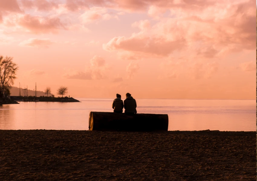 Dos personas sentadas en un gran tronco junto a una tranquila masa de agua al atardecer, tal vez reflexionando sobre la ansiedad en la relación, mientras las nubes flotan sobre sus cabezas y los árboles lejanos bordean la tranquila escena.