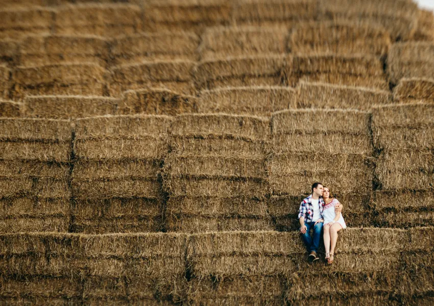 Una pareja se sienta muy unida sobre balas de heno apiladas, rodeada de grandes hileras de heno en un entorno al aire libre, compartiendo un momento de tranquilidad lejos de la ansiedad en la relación.