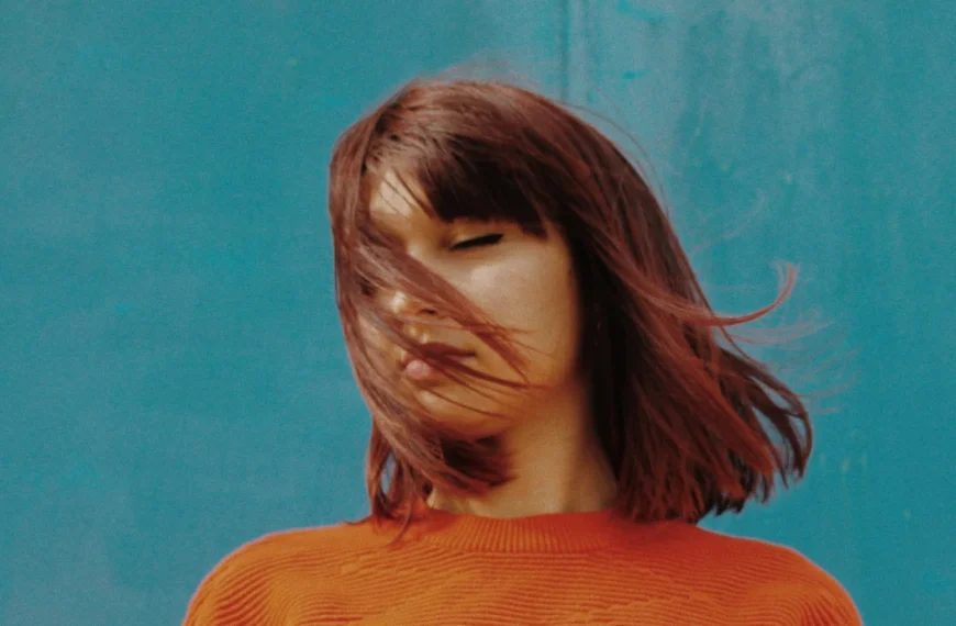 Una persona con el pelo castaño hasta la barbilla que lleva un jersey naranja está de pie sobre un fondo azul, con el pelo cubriéndole parcialmente la cara, como perdida en autoestima frases de reflexión.