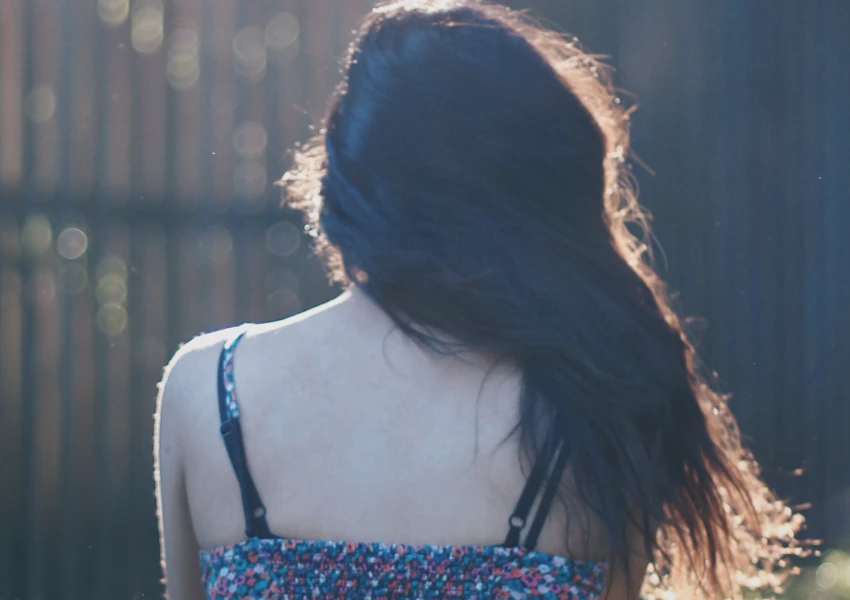 Una persona de pelo largo y oscuro está de pie al aire libre, mirando hacia otro lado, con un top de estampado floral y tirantes finos, a la suave luz del sol, evocando el ambiente tranquilo de la autoestima frases de reflexión.