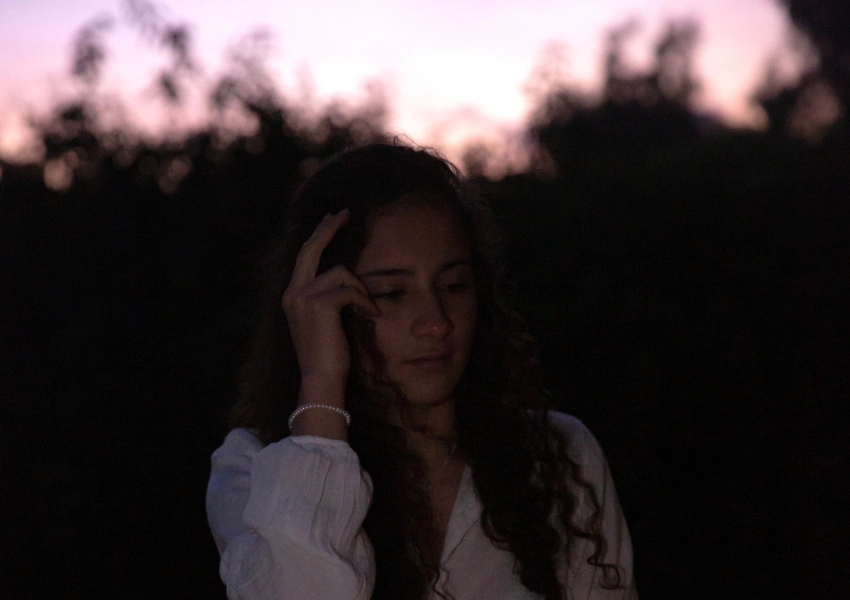 Una mujer joven de pelo largo y rizado está de pie al aire libre al atardecer, tocándose la cara pensativamente. Vestida con una camisa blanca de manga larga, parece reflexionar sobre los estres buenos y malos y su impacto en su bienestar.