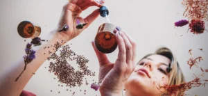 Una persona sostiene un frasco cuentagotas, rodeado de semillas flotantes, flores secas y hierbas sobre un fondo claro, que evoca la relajación y la esencia calmante de la aromaterapia.