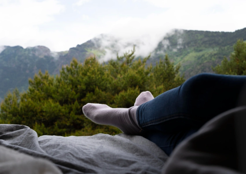 Una persona en calcetines y vaqueros se relaja con las piernas estiradas en una cama, disfrutando de los verdes árboles y las montañas neblinosas al otro lado de la ventana: perfecto para relajarse en 5 minutos.